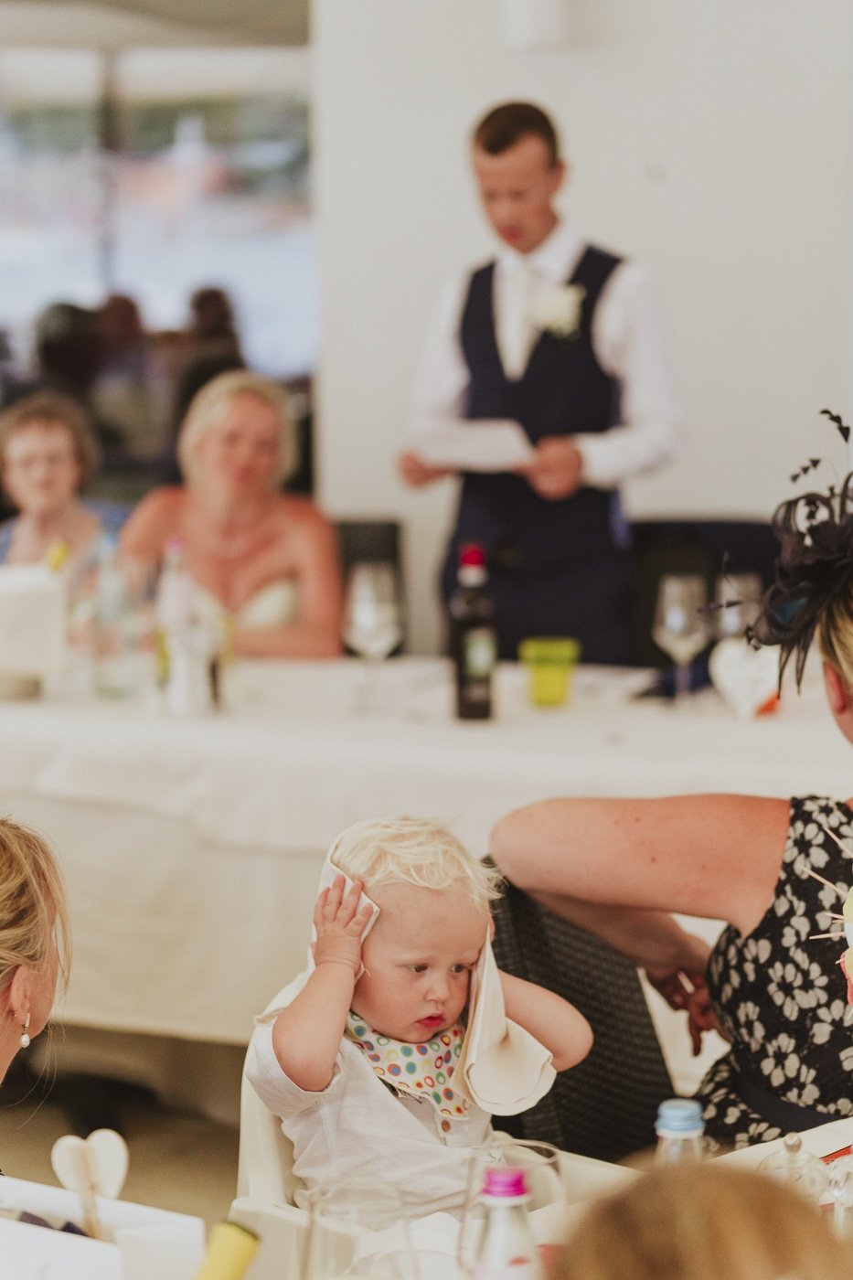blonde haired baby with hands over ears with speeches happening in background by Joshua Wyborn