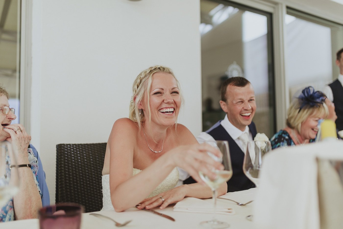 Bride with braids in her hair and a glass of wine in her hand laughs along with husband and guests by Joshua Wyborn
