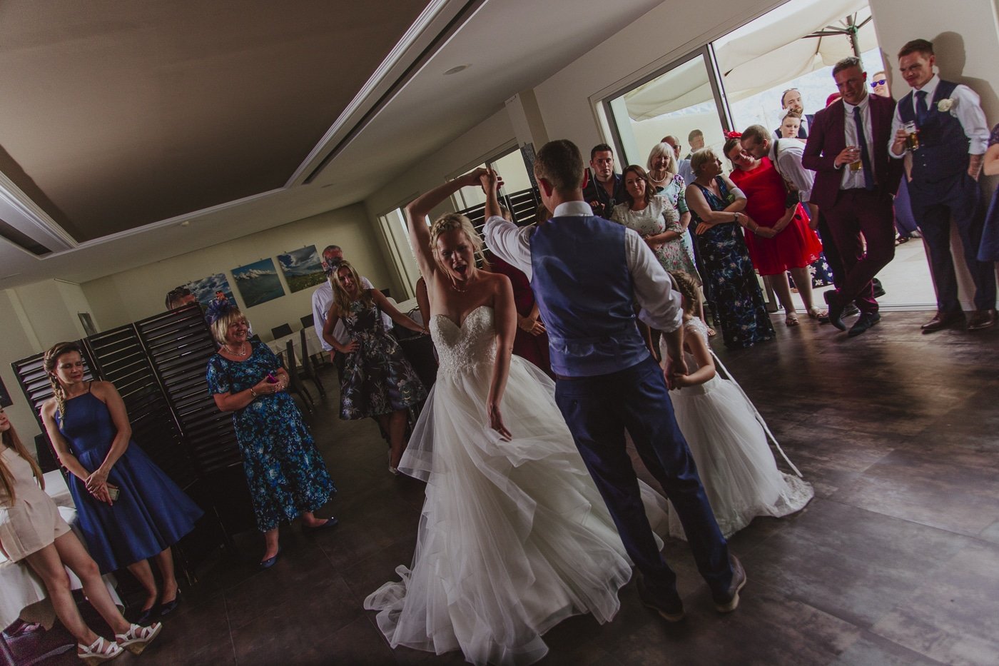 Happy bride spinning on the dance floor by her husband and small bridesmaid whilst wedding guests look on by Joshua Wyborn