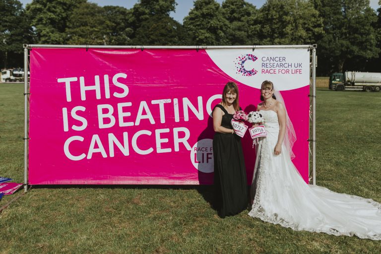 Two brides participating in the Race For Life, standing proudly in front of a banner that says this is beating cancer.