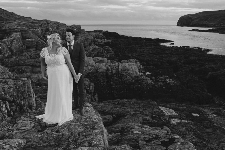 A bride and groom standing on a rocky cliff overlooking The Sound.