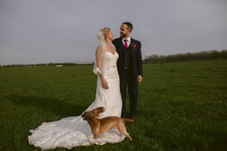 A man and woman in wedding attire pose with a dog in the Three Hills Barn field.