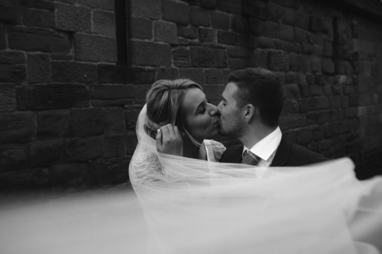 A bride and groom kiss in front of Scotby Church's brick wall.