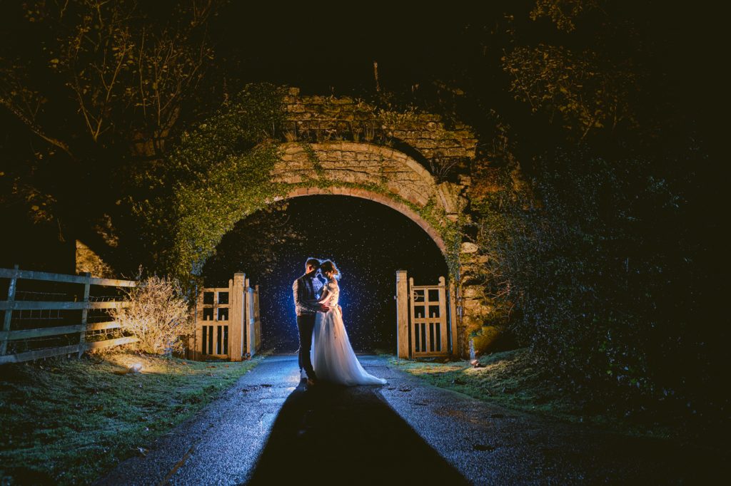A bride and groom standing in front of the Lanercost Priory archway at night.