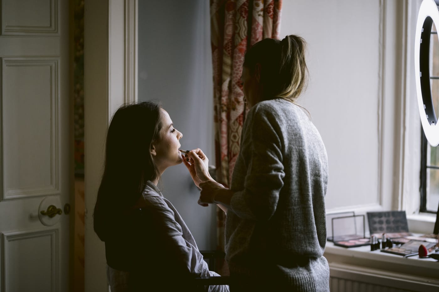 A woman is getting her makeup done in front of a Askham window.
