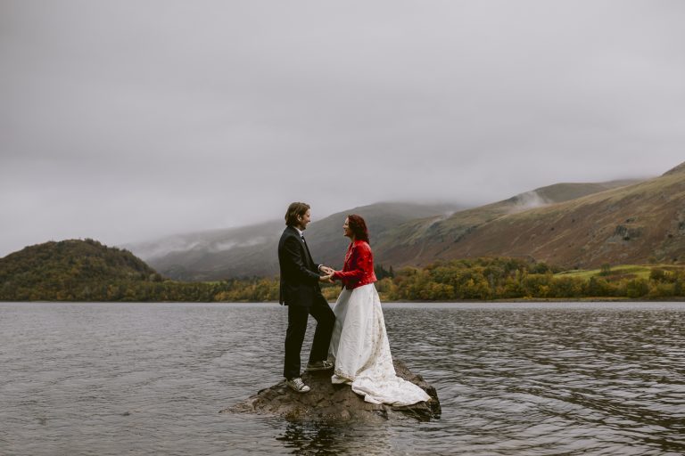 A bride and groom celebrating their wedding anniversary standing on a rock in the lake.