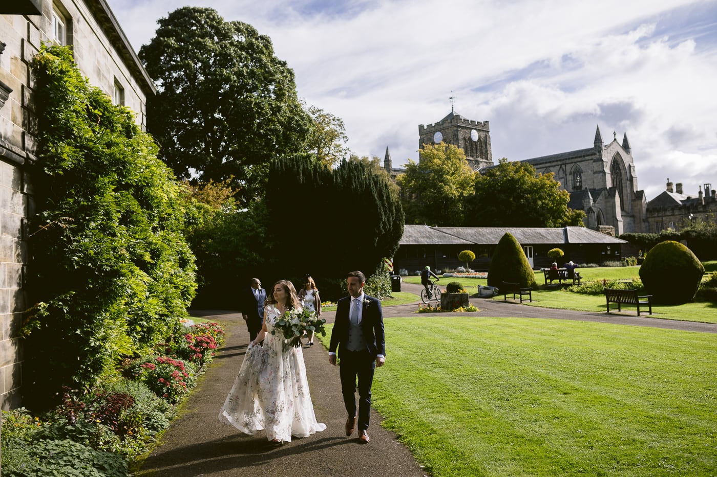 A Hexham House wedding with a bride and groom walking through a garden with a castle in the background.