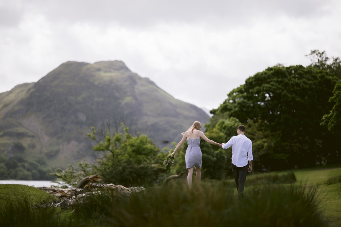 A couple walking through a field with mountains in the background in the Lake District, for their Civil Partnership celebration.