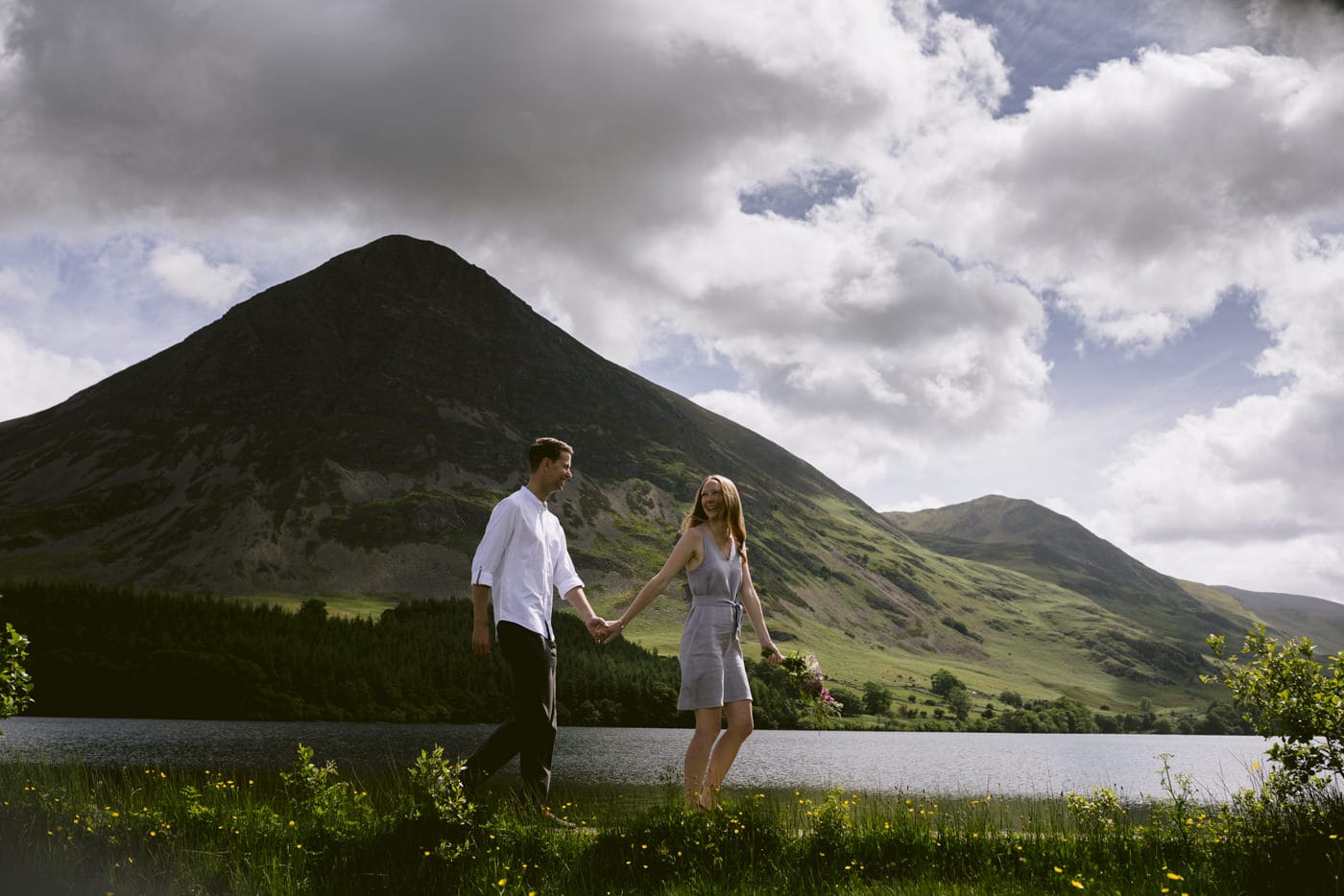 A couple embracing and strolling along the serene shores of a picturesque lake, with majestic mountains framing the background.