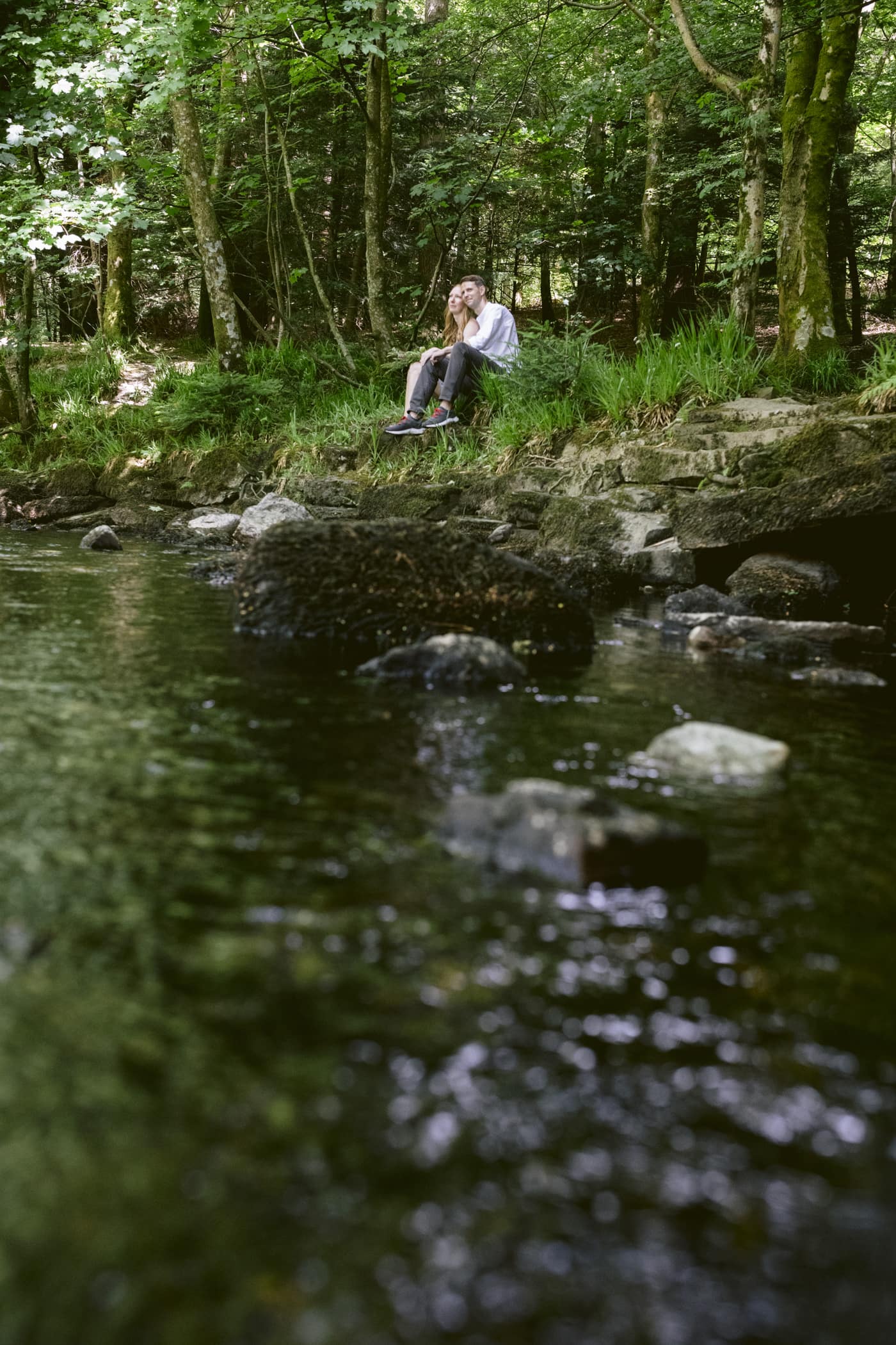 A couple sits near a river in the woods during their Lake District Civil Partnership ceremony.