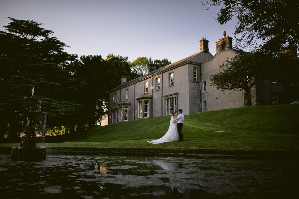 bride and groom outside farlam hall stood by the wedding venues pond