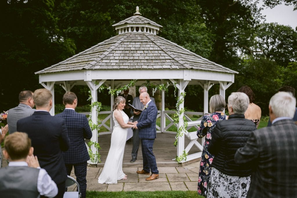 An Outdoor Armathwaite Hall wedding ceremony takes place as the radiant bride and groom exchange vows under a picturesque gazebo.