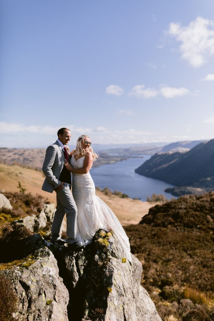 An adventurous bride and groom stand on top of Ullswater Mountain, overlooking the lake after a hike.