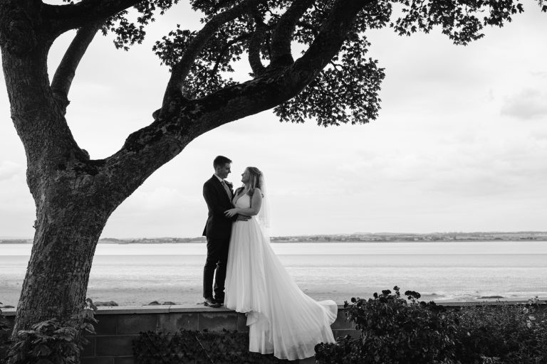 A bride and groom standing under a tree in front of a lake on the Solway Coast.