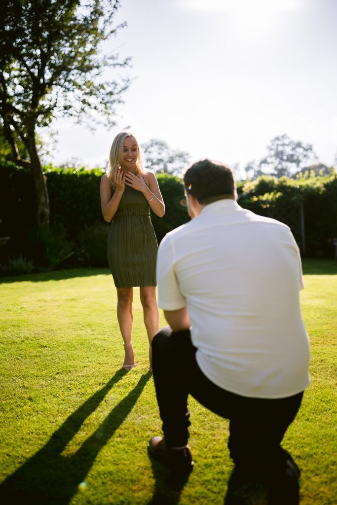 A man kneeling down to propose to a woman in the grass.