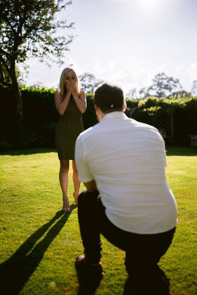A man kneeling on the grass to propose to a woman.