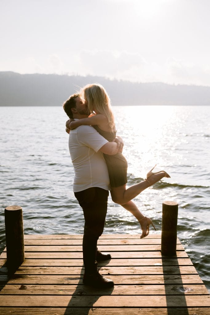 A man and woman hugging on a dock in front of a lake.