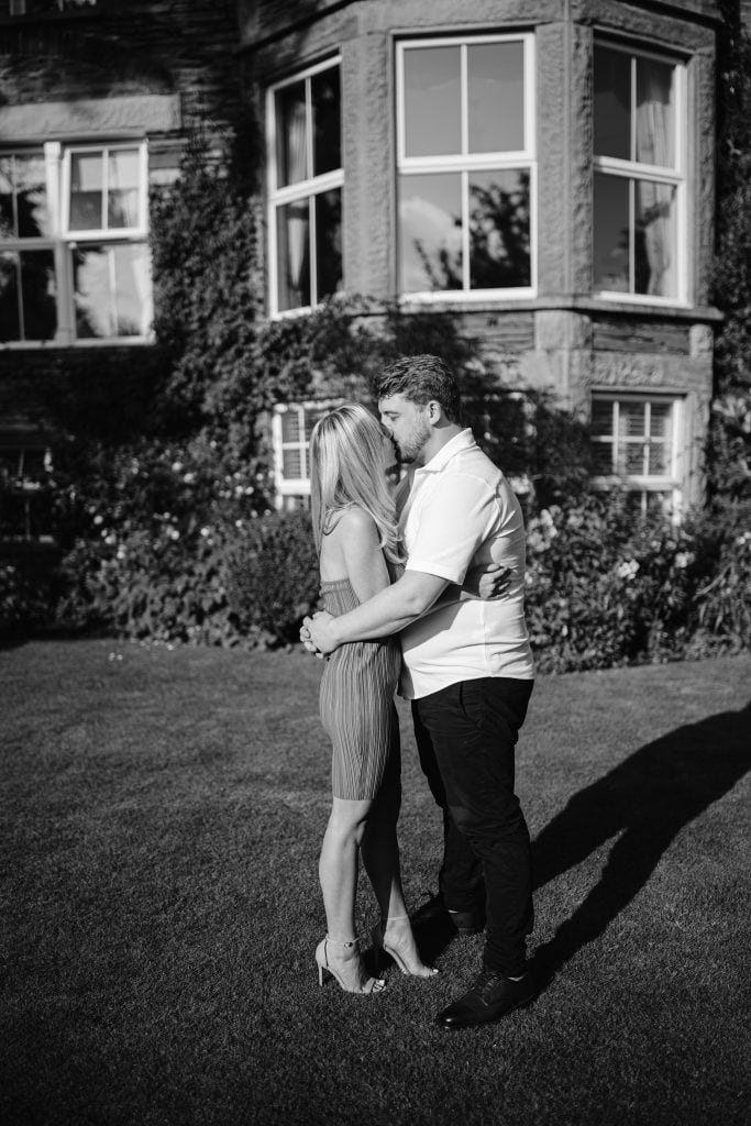 A black and white photo of a couple embracing in front of a large house.