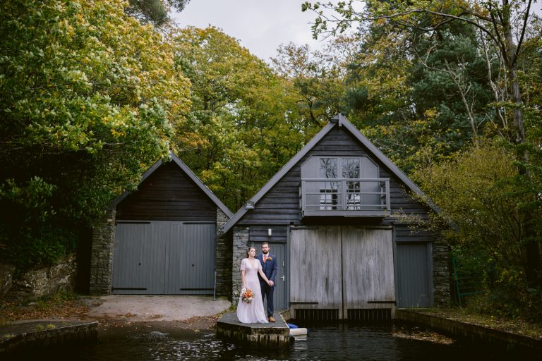 An autumn elopement in the Lake District as the bride and groom stand in front of a boat amidst the enchanting woods.