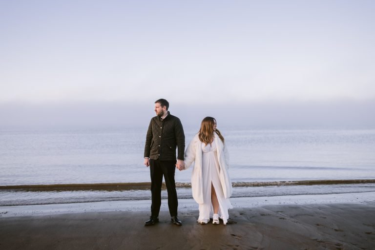 A bride and groom having a romantic seaside portrait session on the beach, holding hands.