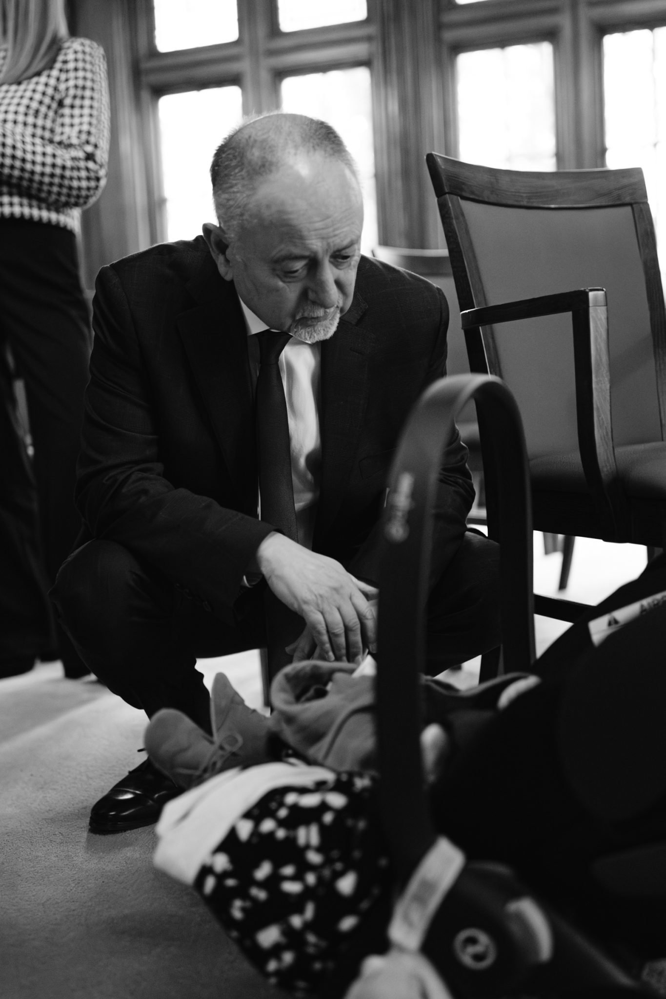 A man in a suit kneels down next to a baby in a car seat outside Lady Gillford's house during the winter.