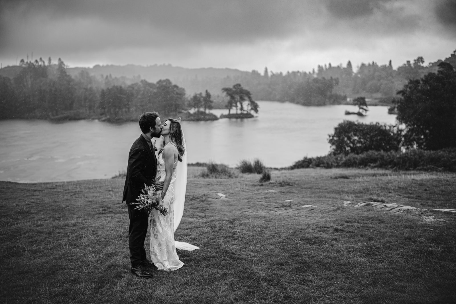 A man and woman sharing a romantic kiss on a grassy hill overlooking the serene water of Yew Tree Farm on a delightful summer day.