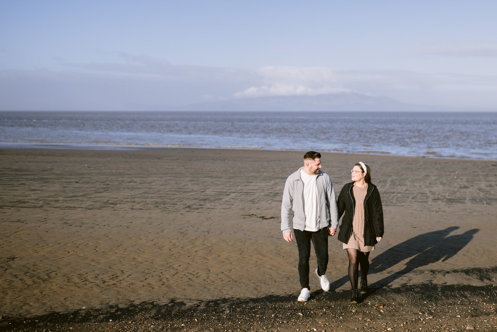 A couple holding hands during their Portrait Session while walking on a sandy beach with a body of water and mountains in the distance.