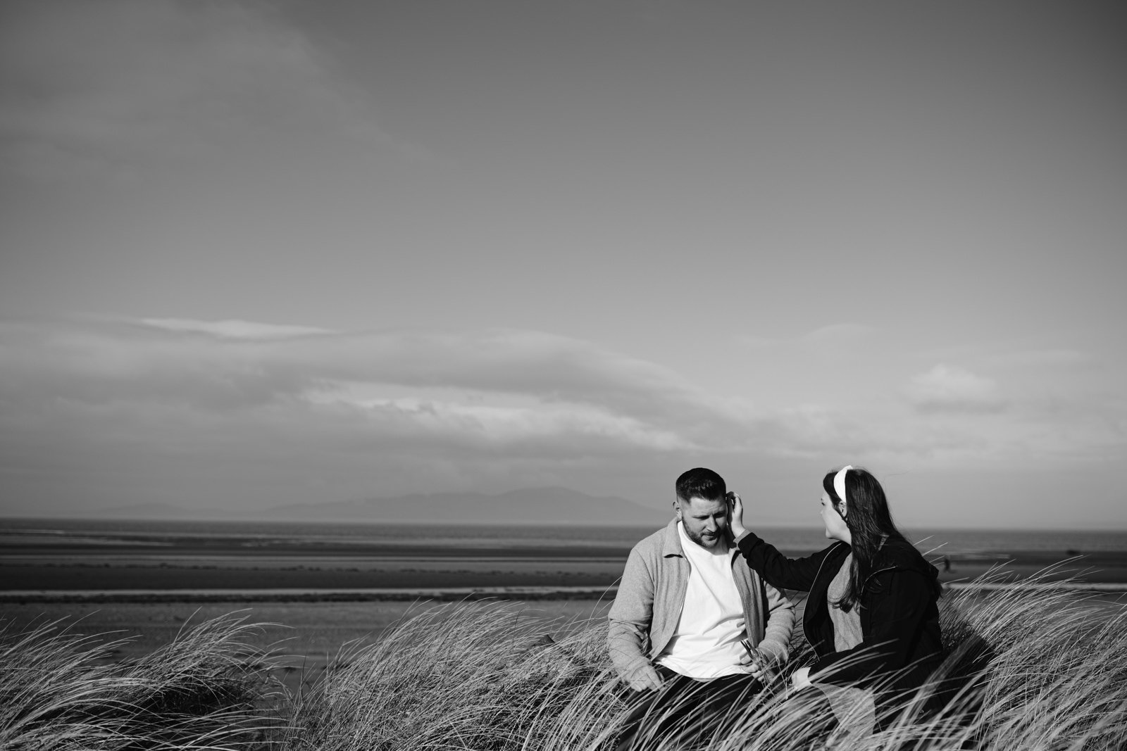 Two people sitting in tall grass, engaging in conversation with a scenic landscape of a lake and waterfalls in the background.
