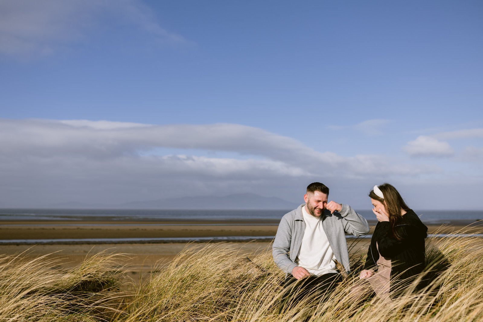 Two people sitting in tall grass by the lakeshore, sharing a conversation under a clear sky during their couple portrait session.
