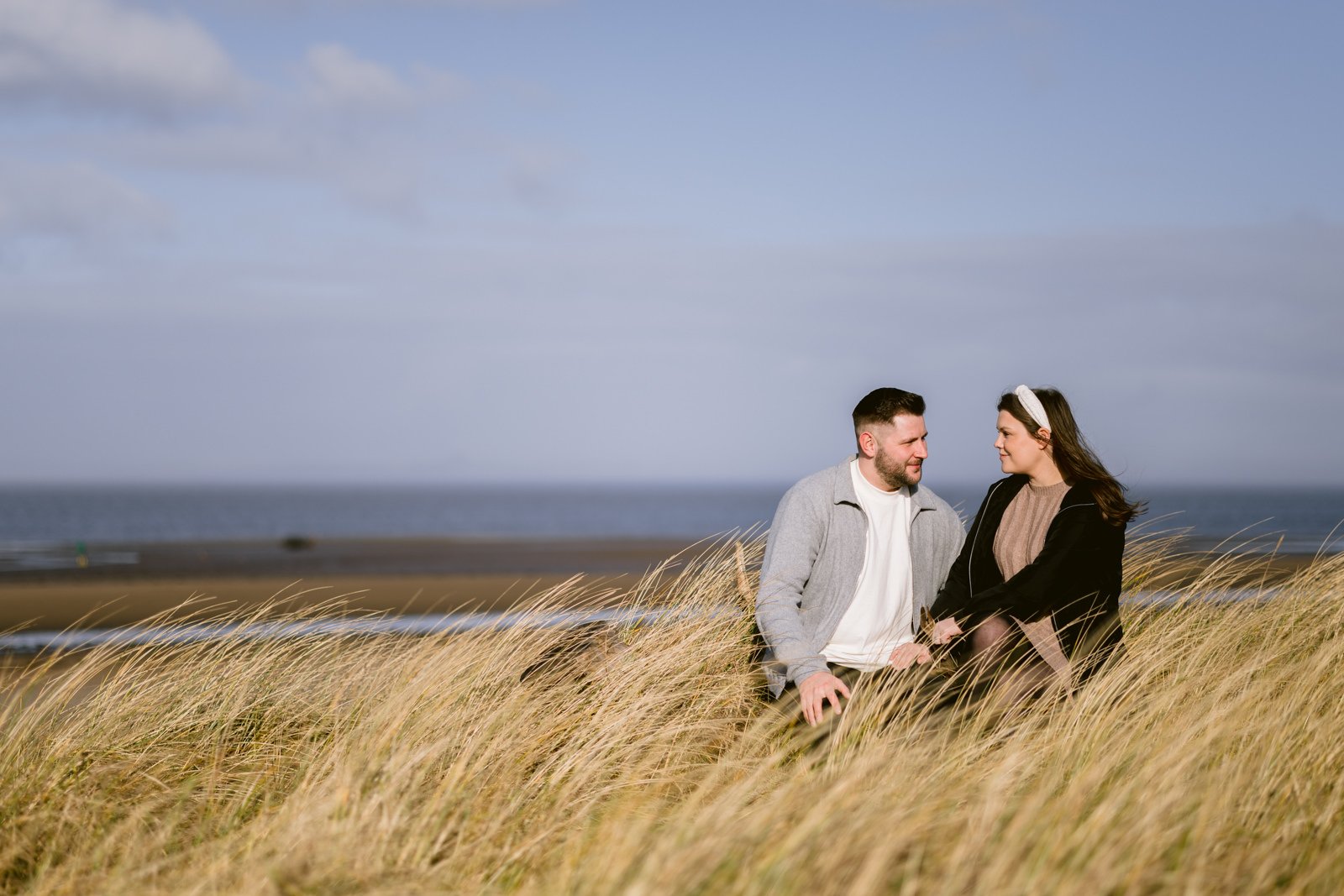A couple sitting in tall grass on a coastal dune, looking at each other during their Portrait Session with the sea in the background.