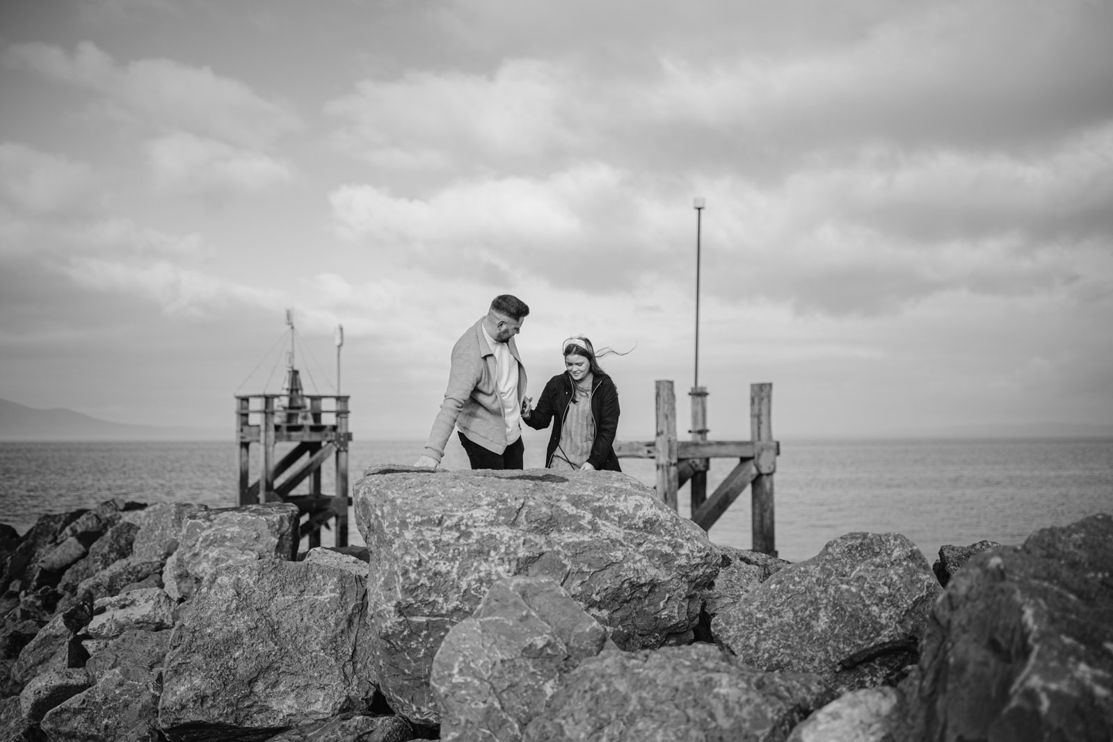 Two people conversing on a rocky shore with a pier in the background, under a cloudy sky during their Portrait Session.