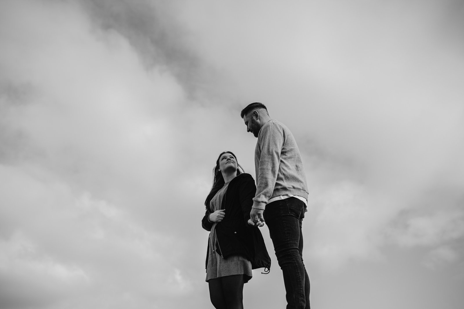Two people standing under a cloudy sky beside a river, one looking up with a hand on their hip, the other glancing down with a smile.
