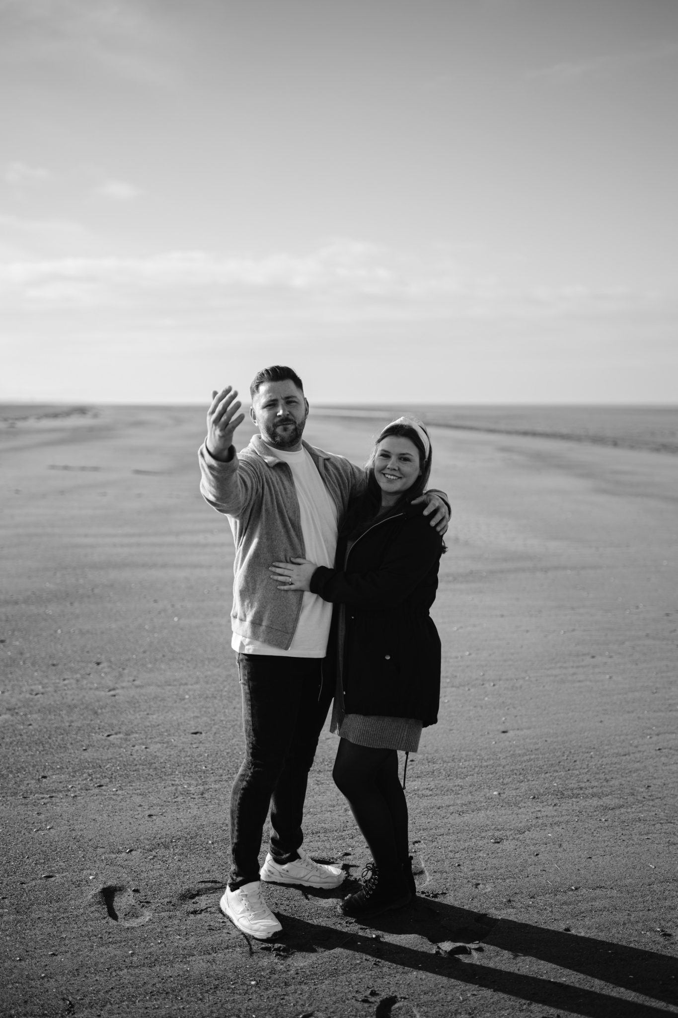 A couple standing on the expansive shore of a lake while smiling, with one person reaching out towards the camera during their portrait session.