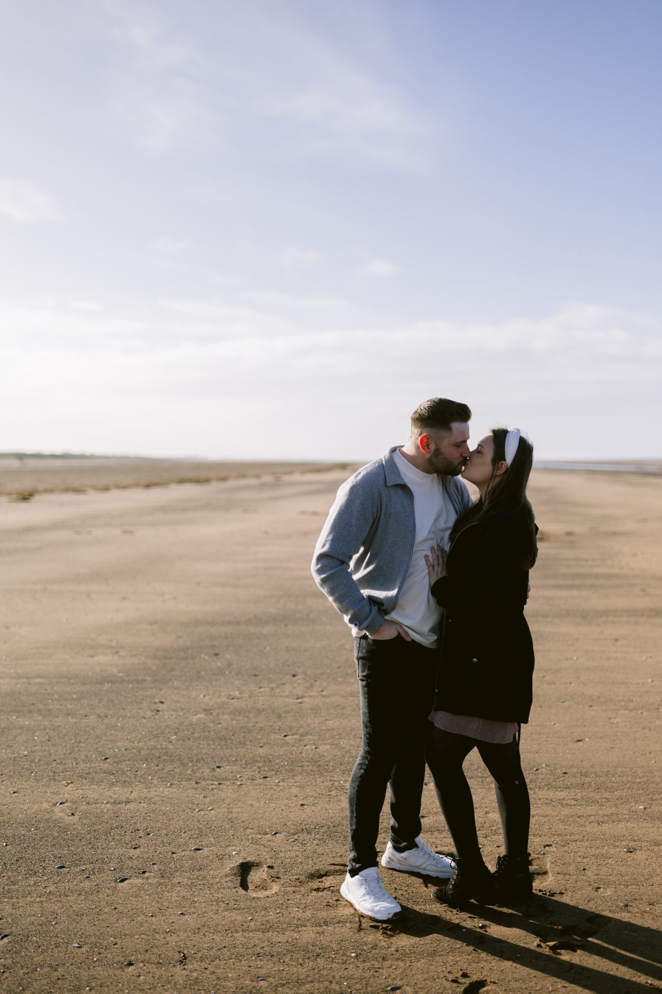 A couple embracing on a sandy beach by a lake under a clear sky during their Portrait Session.