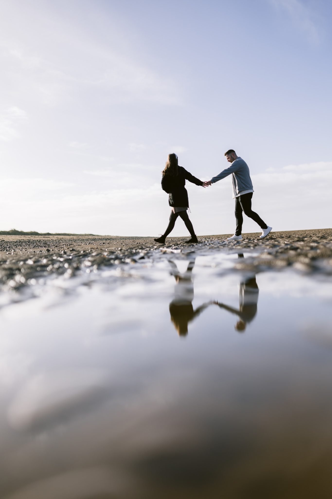 Two people holding hands, with their reflection visible in a water puddle during their couple portrait session.