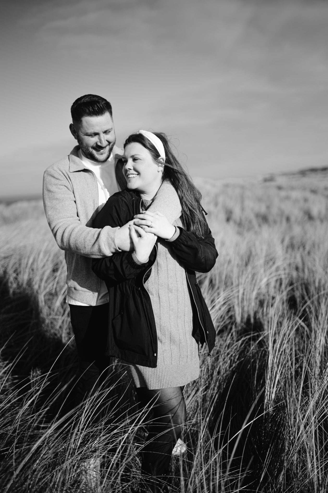 A couple embracing and smiling in an open field with tall grass during their lakeside Portrait Session.