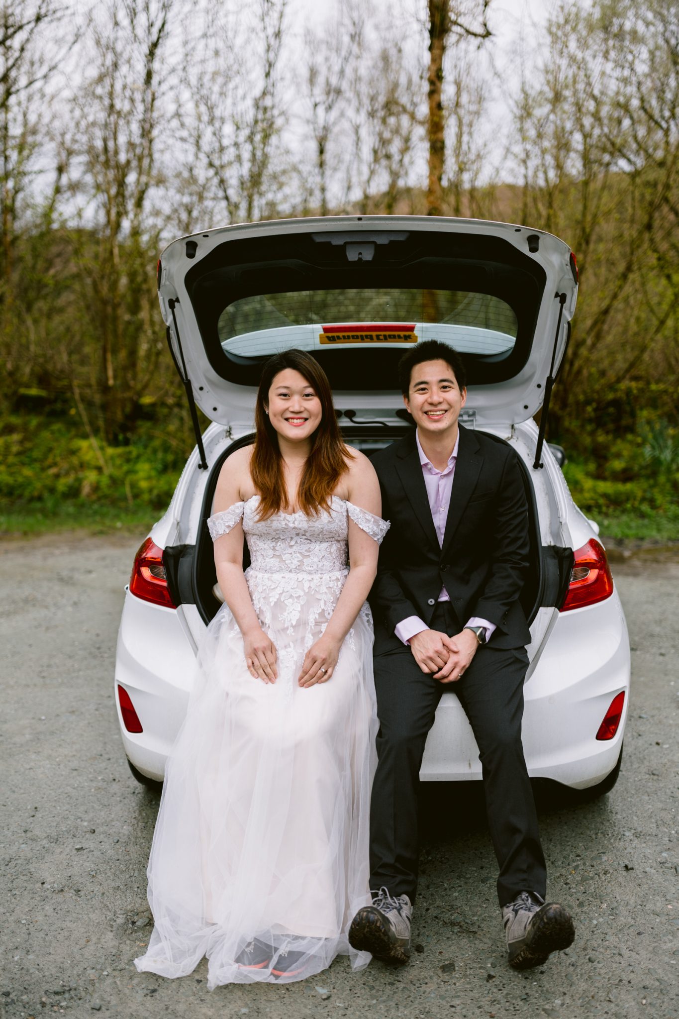 A couple in formal attire celebrating their wedding anniversary, sitting in the trunk of a car with the hatch open, smiling at the camera.