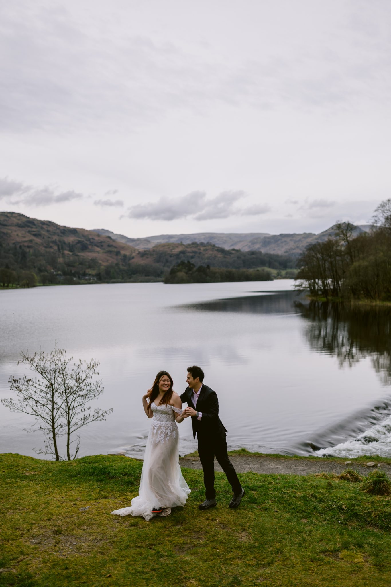 A couple celebrating their wedding anniversary in wedding attire, holding hands by a serene lakeside with hills in the distance.
