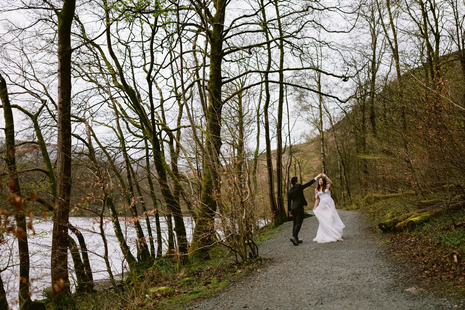 A couple celebrating their wedding anniversary in wedding attire posing on a wooded pathway beside a river.