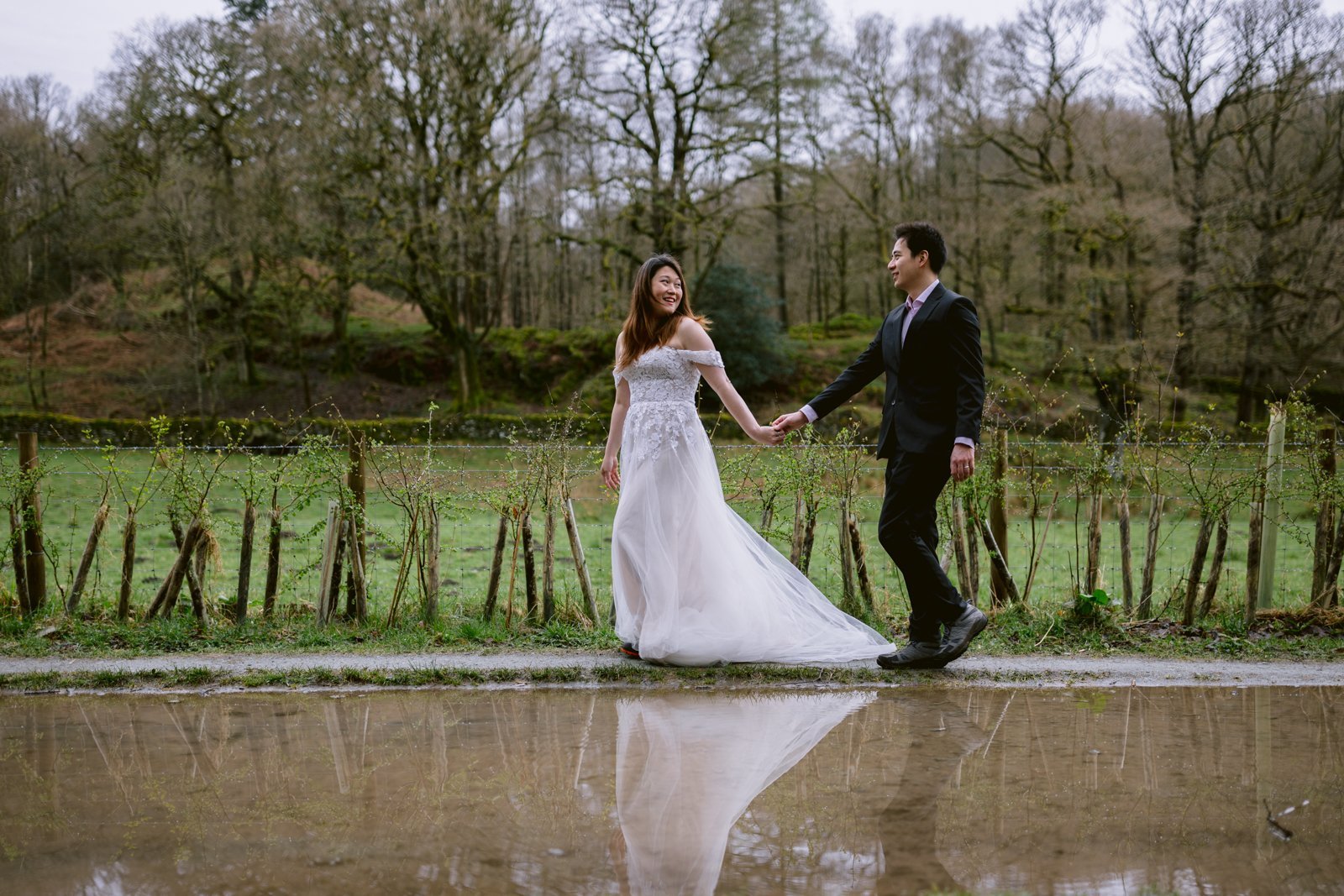 A couple celebrating their wedding anniversary in wedding attire, holding hands near a reflective puddle in a natural setting.