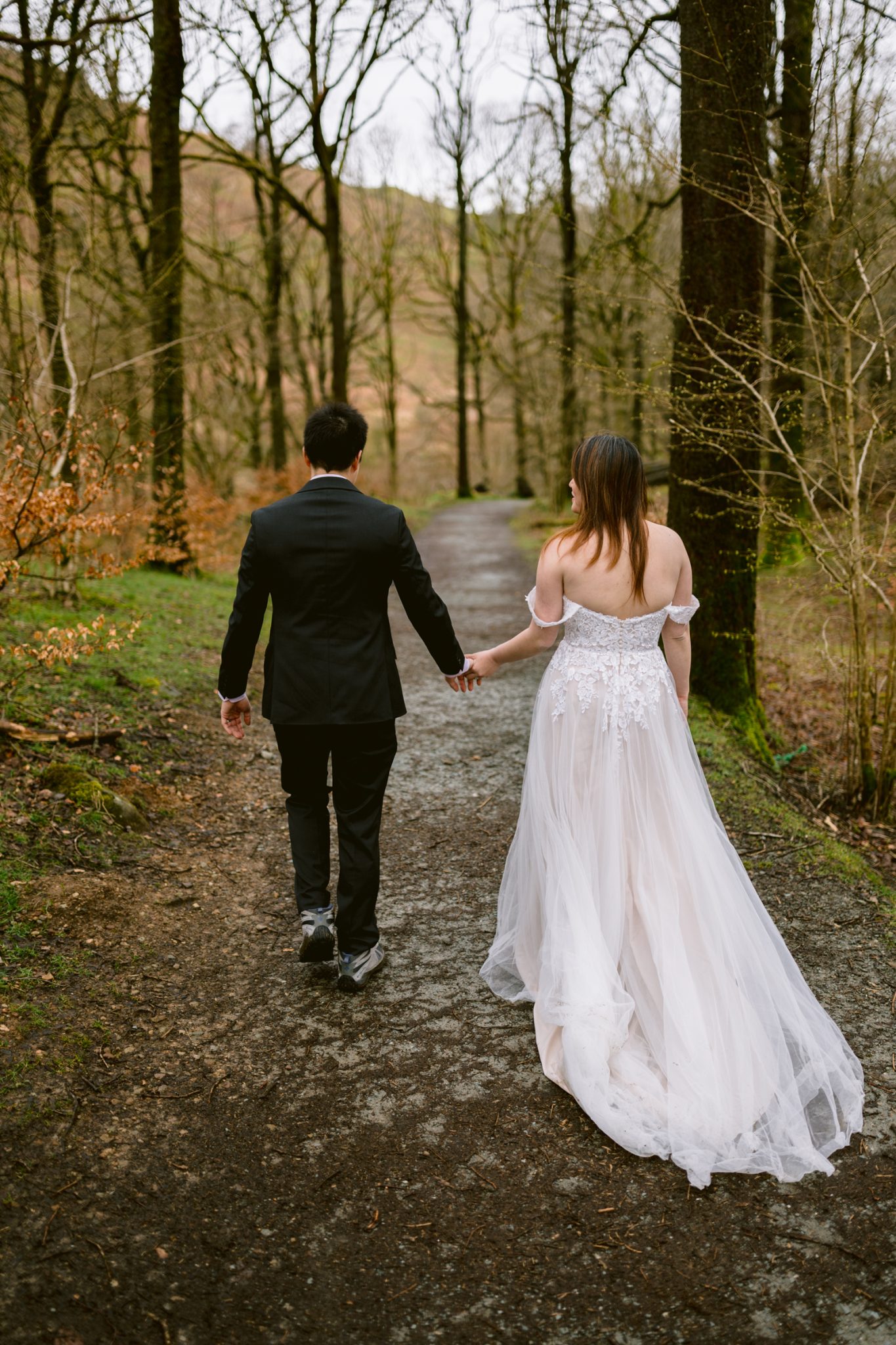 A couple celebrating their wedding anniversary in wedding attire, holding hands and walking down a woodland path.
