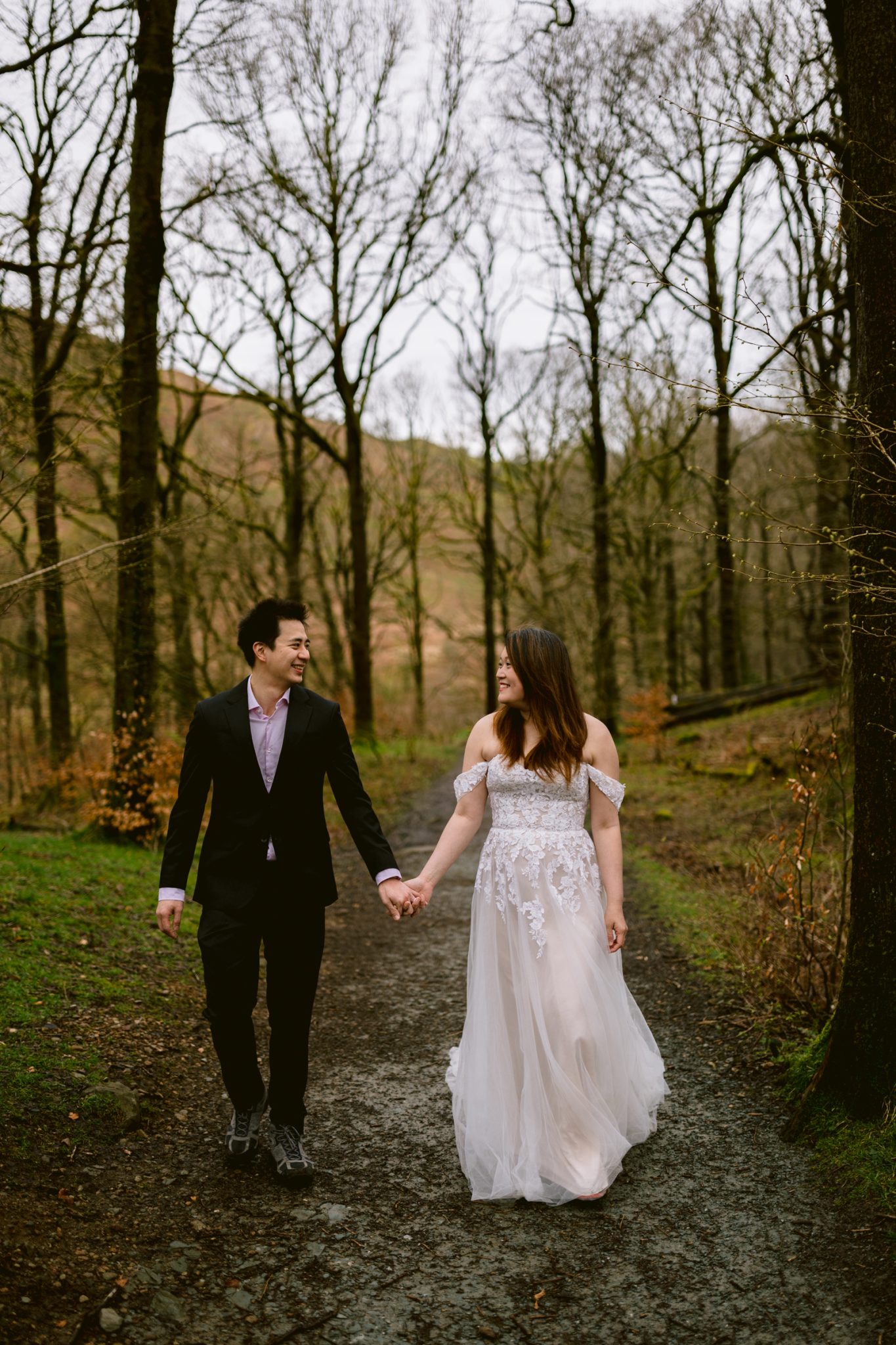 A couple holding hands and walking down a woodland path, dressed in formal attire, celebrating their wedding anniversary.