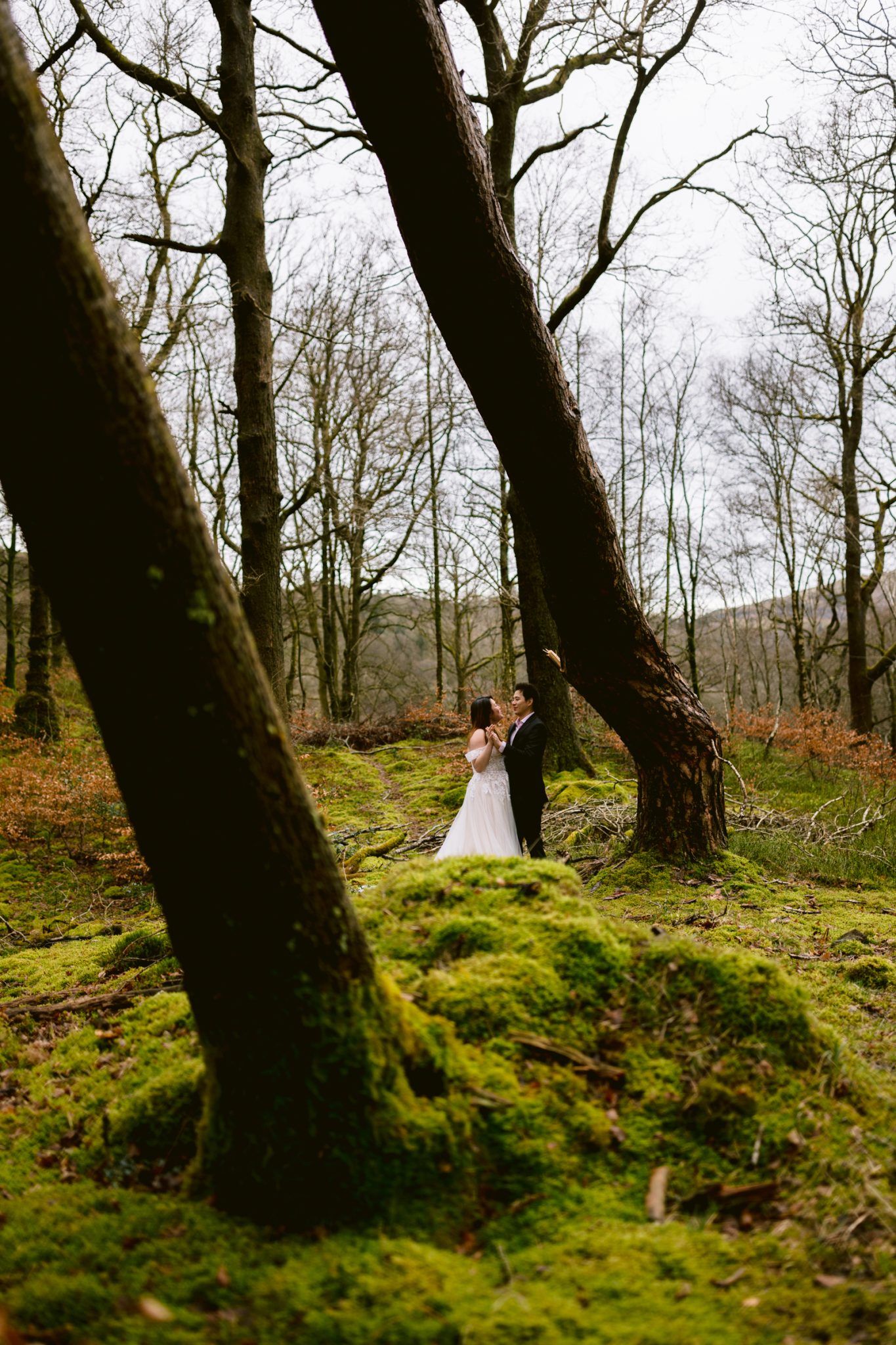 A couple celebrating their wedding anniversary in wedding attire, sharing a moment in a forest with moss-covered ground.