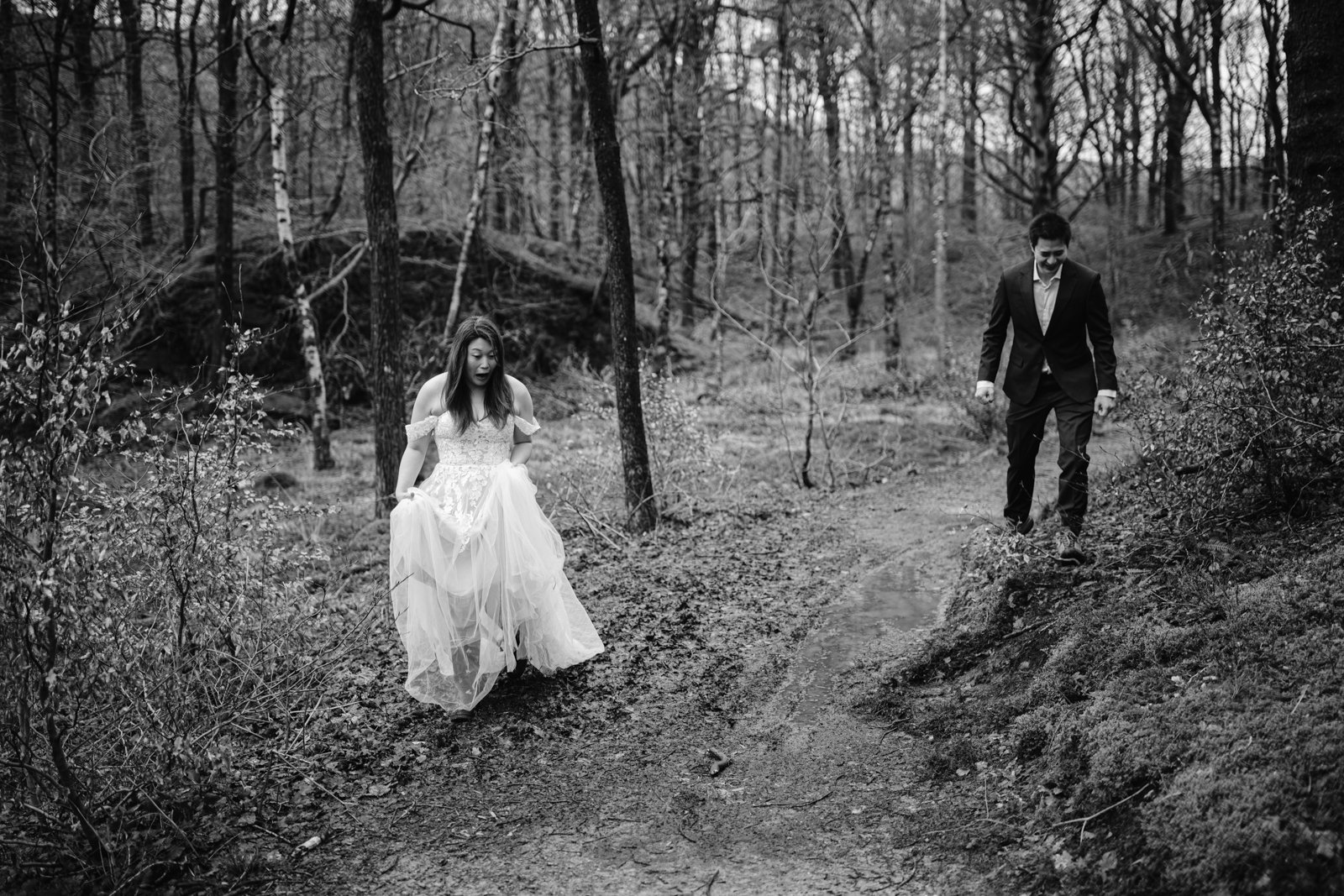 A bride sitting on a dirt path in a forest while a groom walks towards her to celebrate their wedding anniversary.
