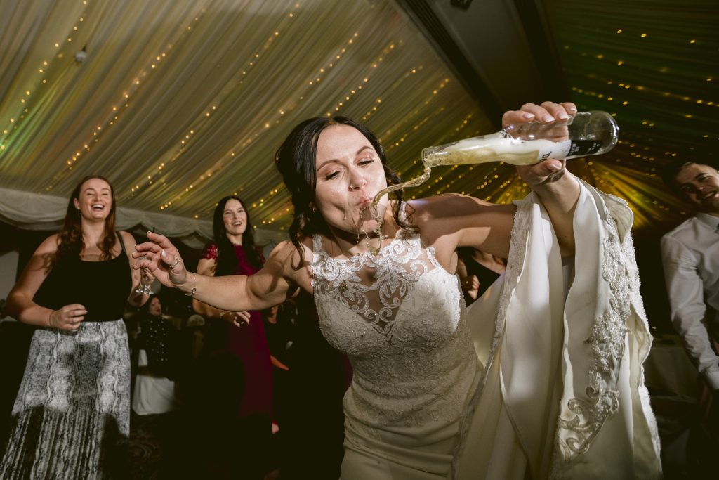 A bride in a white dress is spilling a drink while pouring it into her mouth at an authentic wedding reception, surrounded by guests in a tent adorned with string lights.