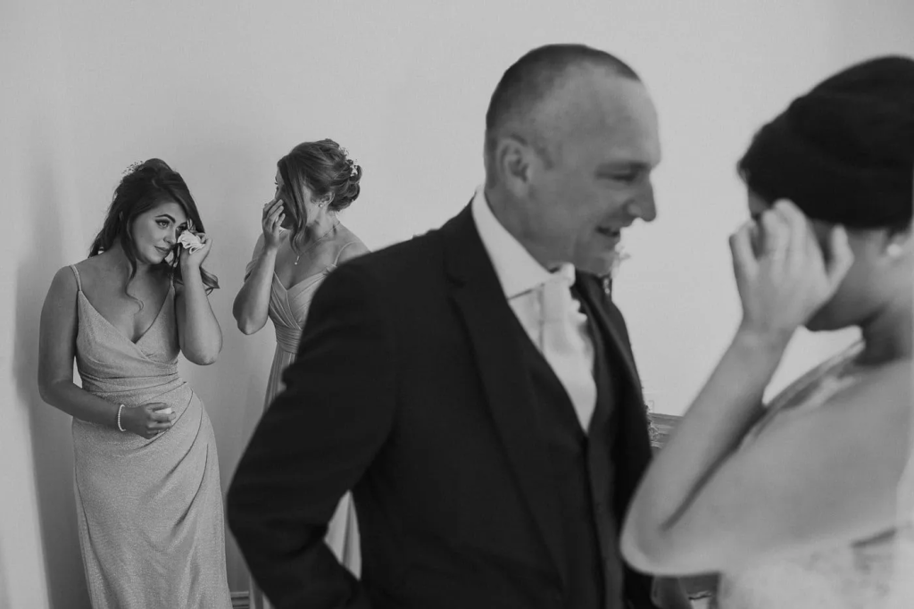 A bride and groom stand together while two bridesmaids in the background wipe their tears, all dressed formally. This scene epitomizes authentic weddings, capturing perfect pictures full of genuine emotion.
