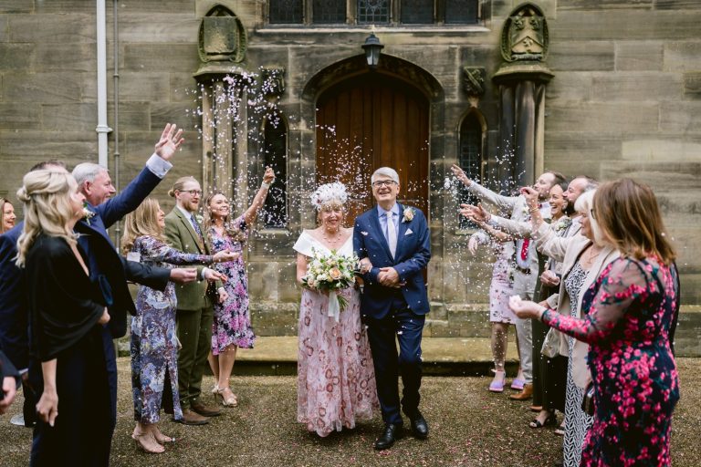 A joyful older couple walks through a shower of confetti thrown by guests outside a Carlisle church, smiling broadly.