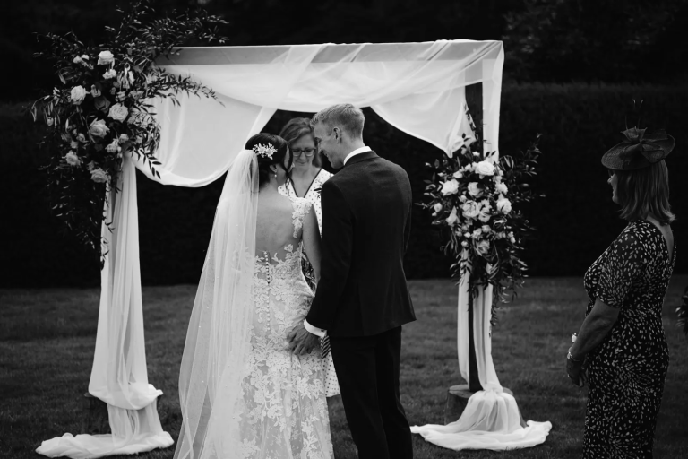 A bride and groom stand facing each other under a floral arch during an outdoor wedding ceremony, creating a picture-perfect moment, with an officiant and a woman standing to the side observing.