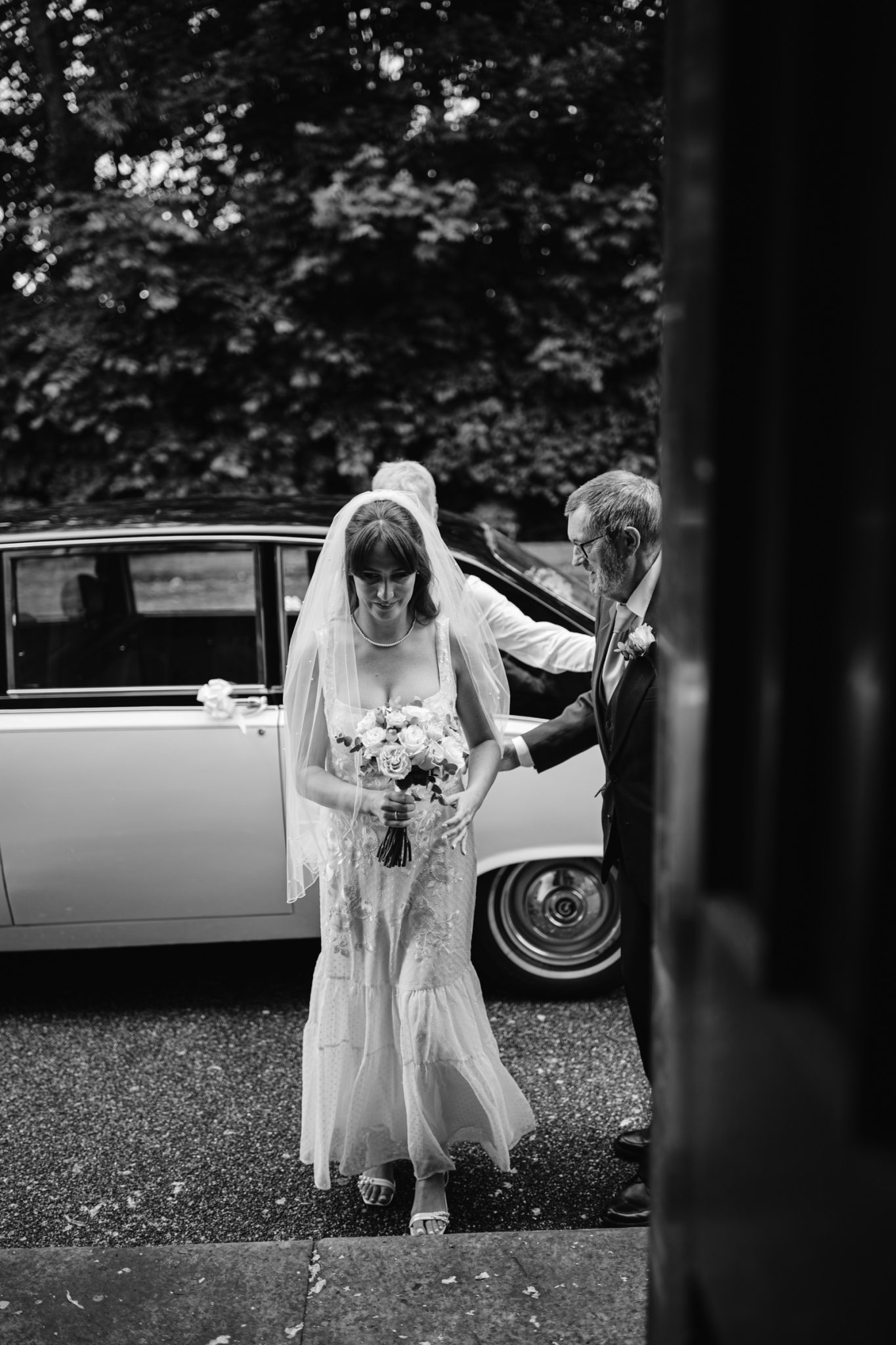 A bride holding a bouquet steps out of a car assisted by an older man in a suit at the Carlisle Registry Office. Another man in a suit opens the car door. Trees and greenery are seen in the background.