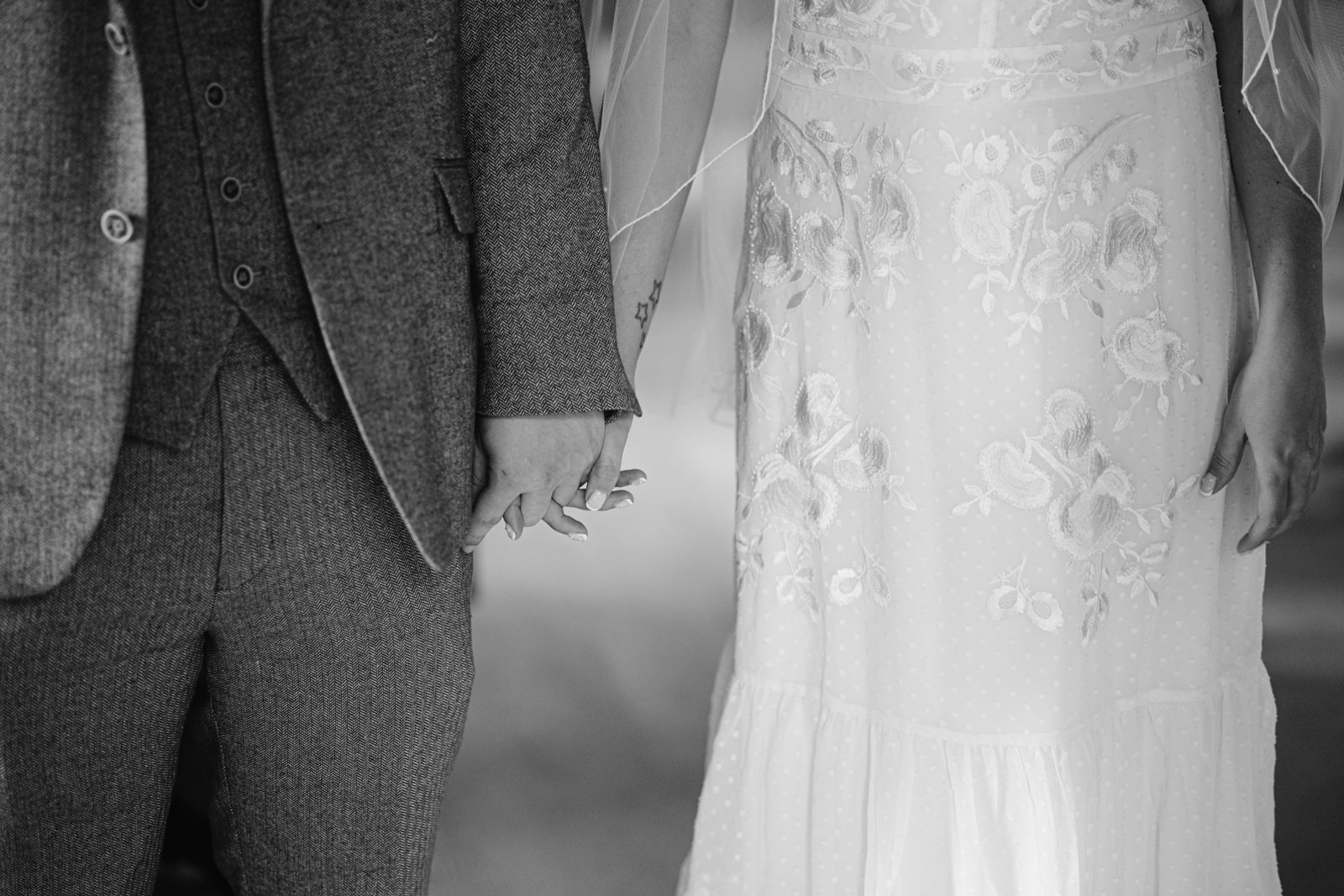 A black-and-white photo of a couple holding hands, taken just outside Carlisle Registry Office. The man wears a suit, and the woman wears a dress with floral embroidery. Their faces are not visible.